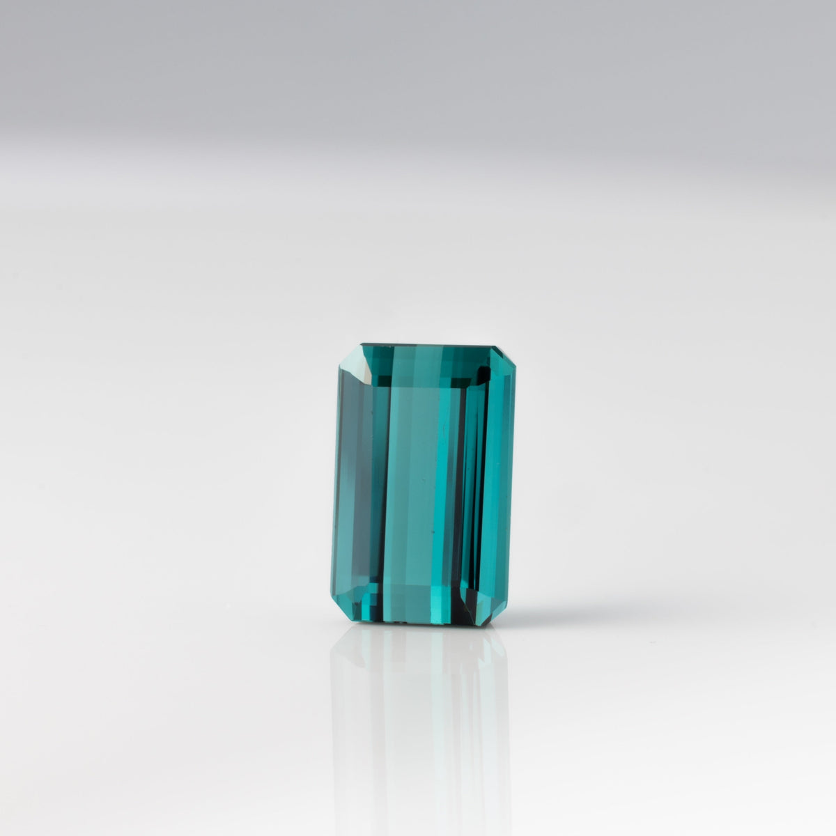 A single rectangular faceted gemstone with a vibrant teal-blue color, standing upright on a reflective white surface against a light gray background.
